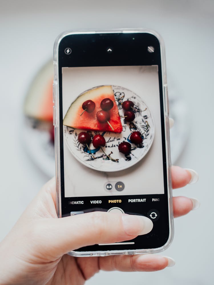 Hand Of A Woman Taking A Smart Phone Photo Of Cherries And A Watermelon Slice On A Plate