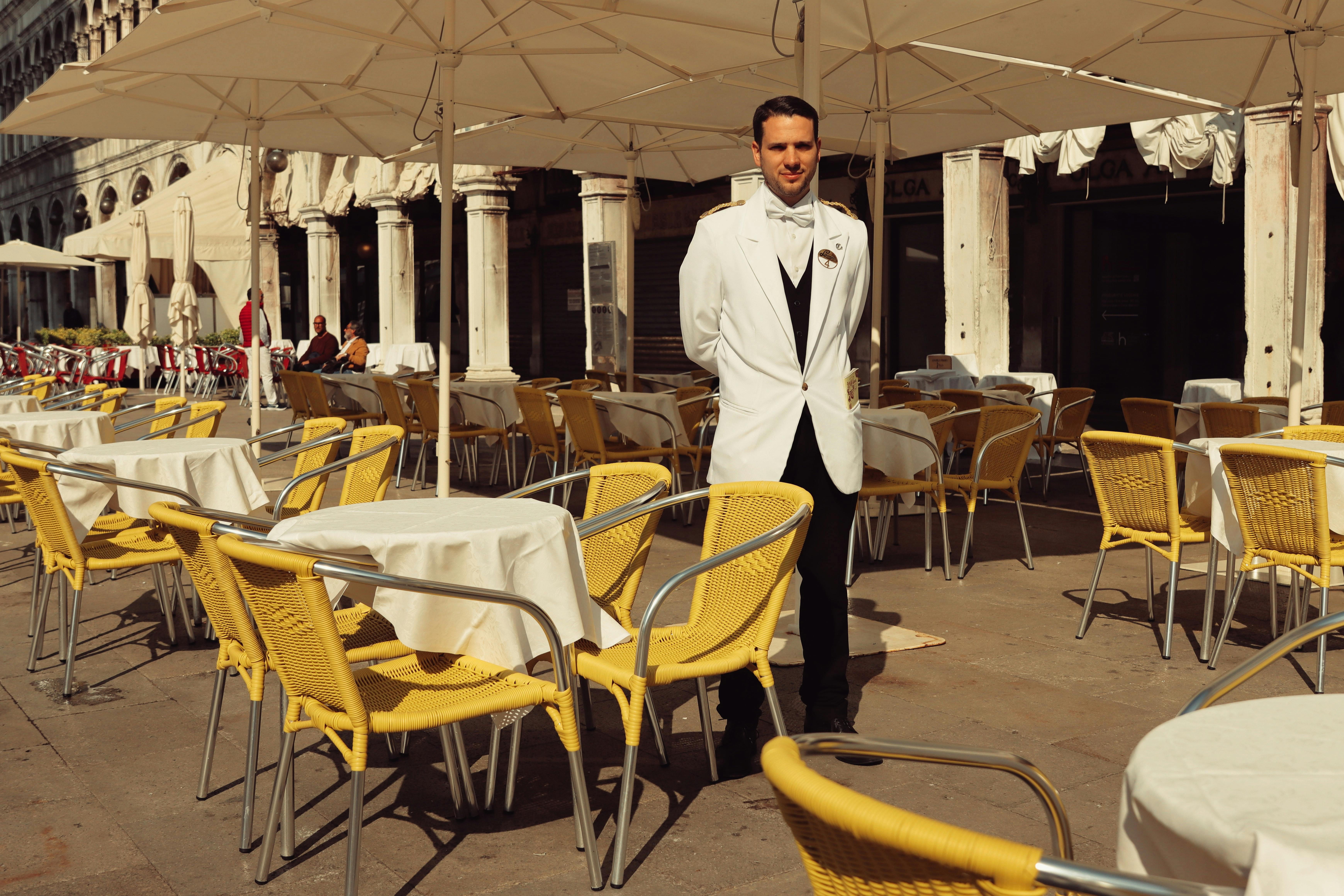 A waiter in a white suit stands amidst sunlit tables in a Venice café.
