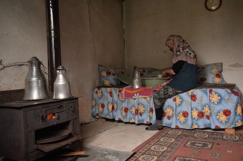 Free Elderly woman in traditional attire preparing food on a couch in a rustic Turkish home with a wood stove. Stock Photo