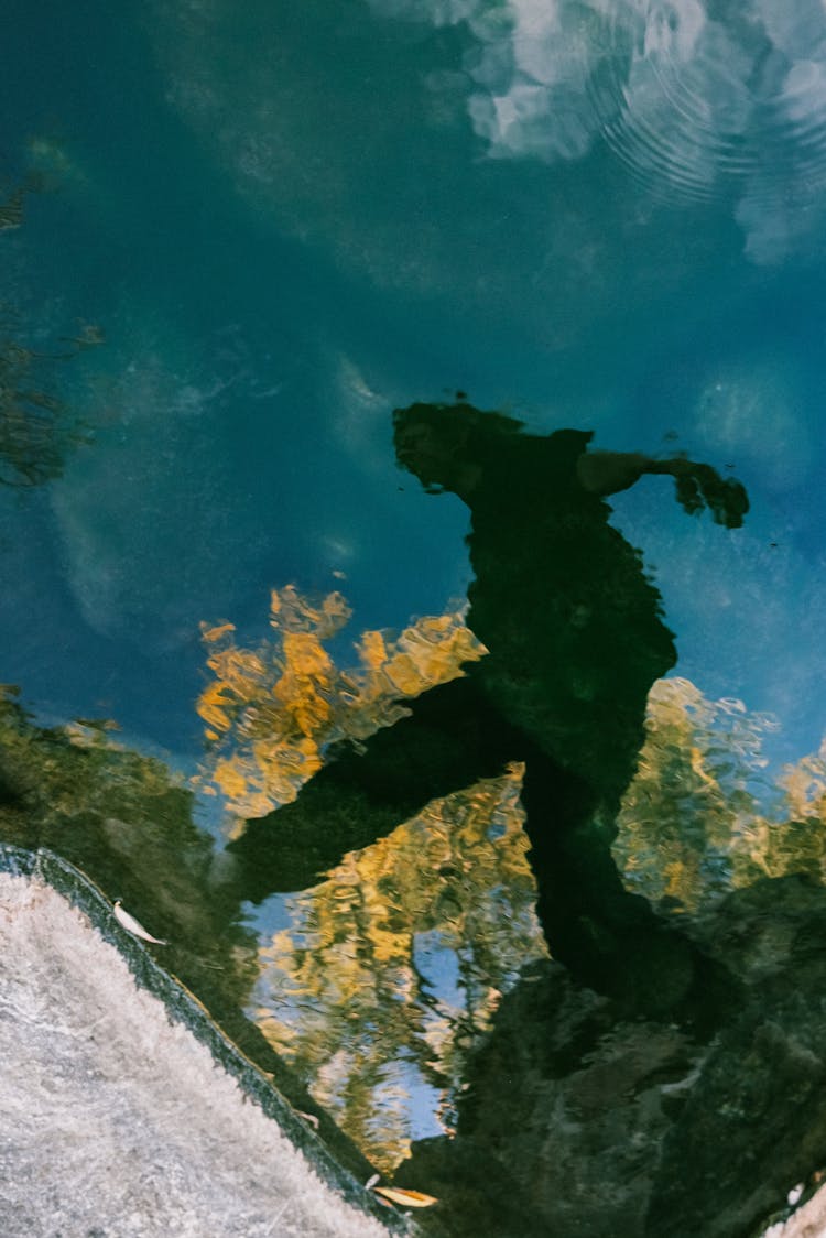 Silhouetted Reflection Of A Woman Jumping On The Rocks In A River Water Surface 