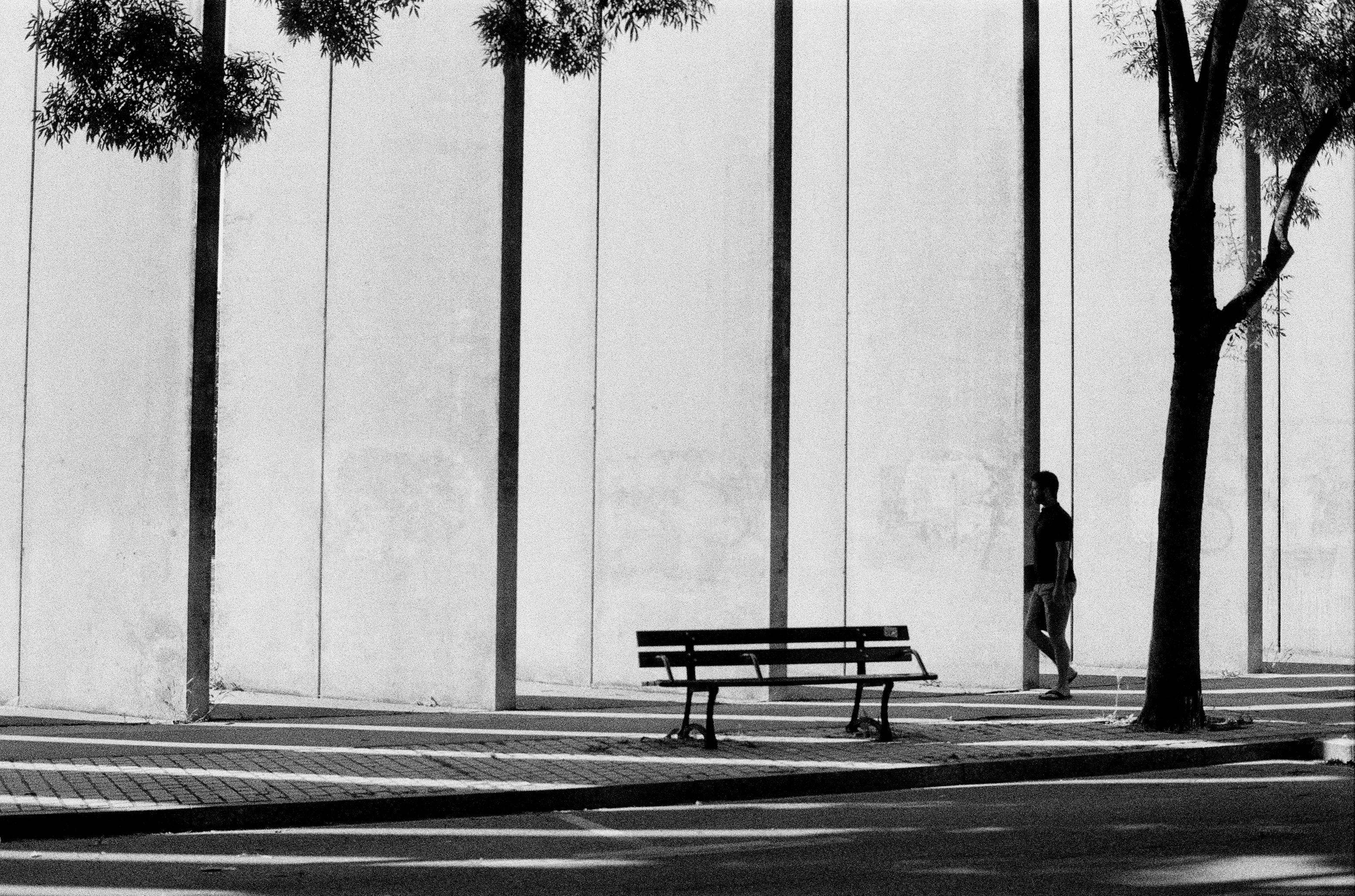 A black and white photo of a man walking past a bench on a sunny day in Nancy, France.