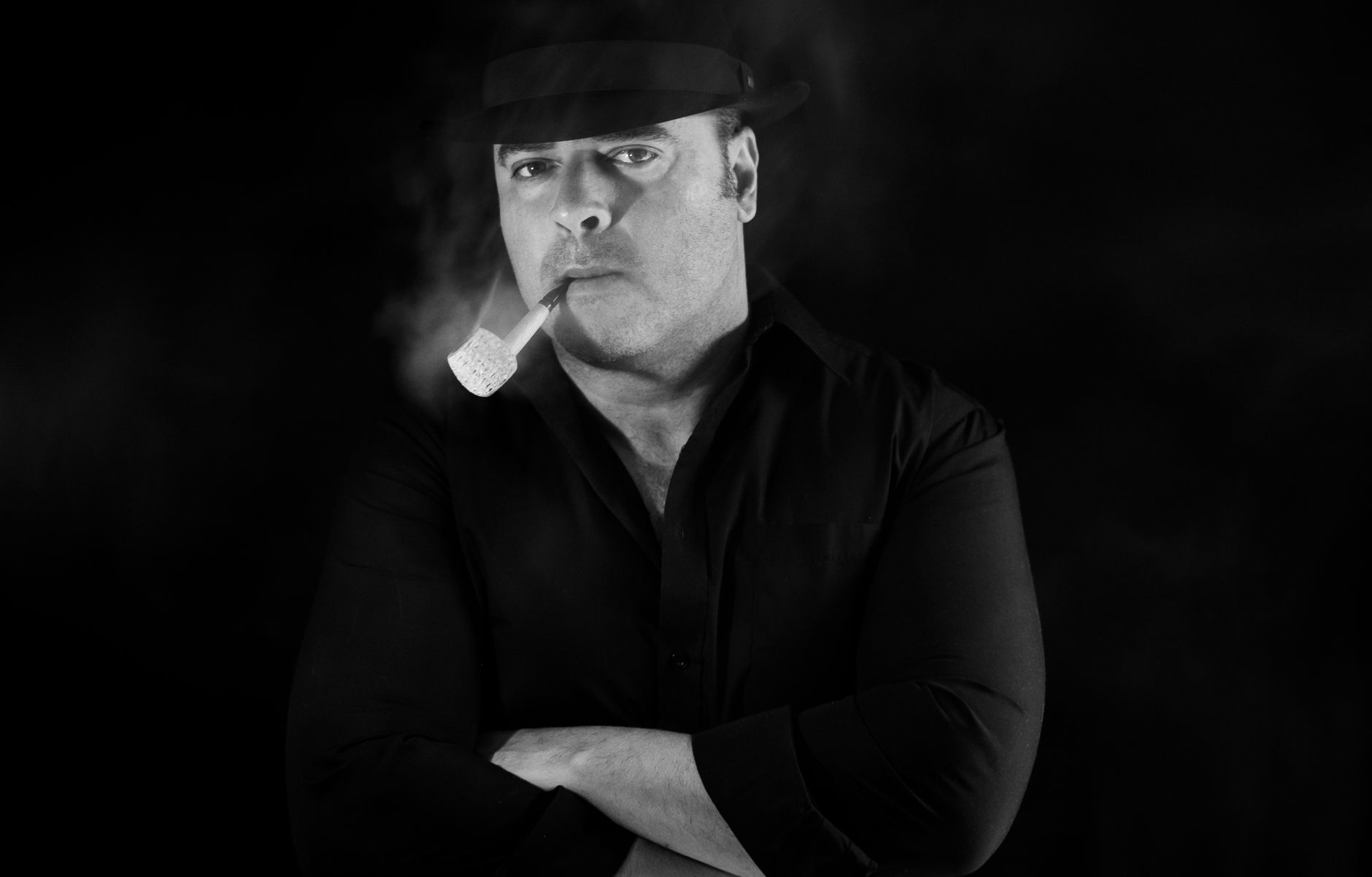 Dramatic black and white portrait of a man in a hat smoking a pipe set against a dark background.