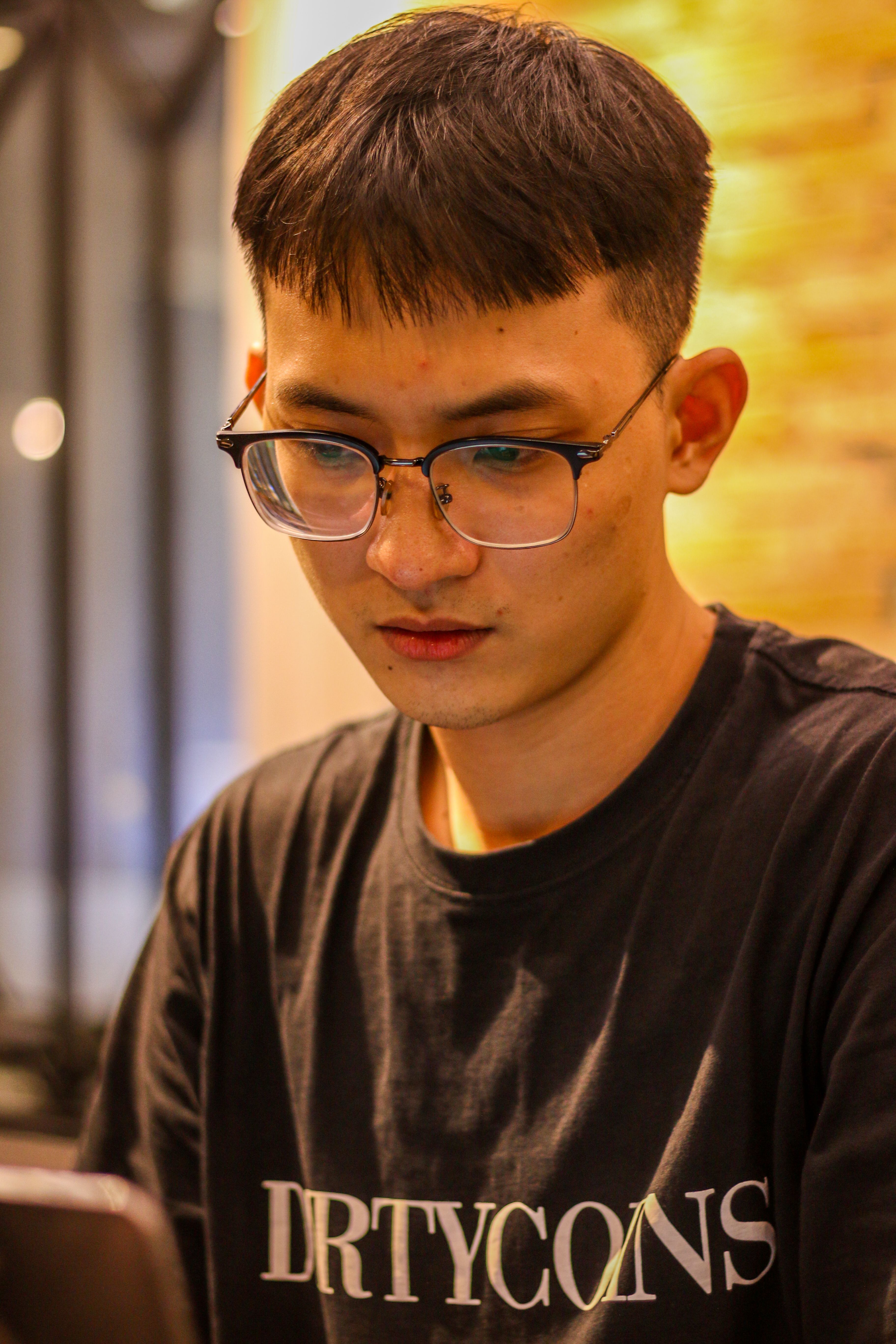 Man with glasses and short hair working focusfully on his laptop indoors, wearing a t-shirt.