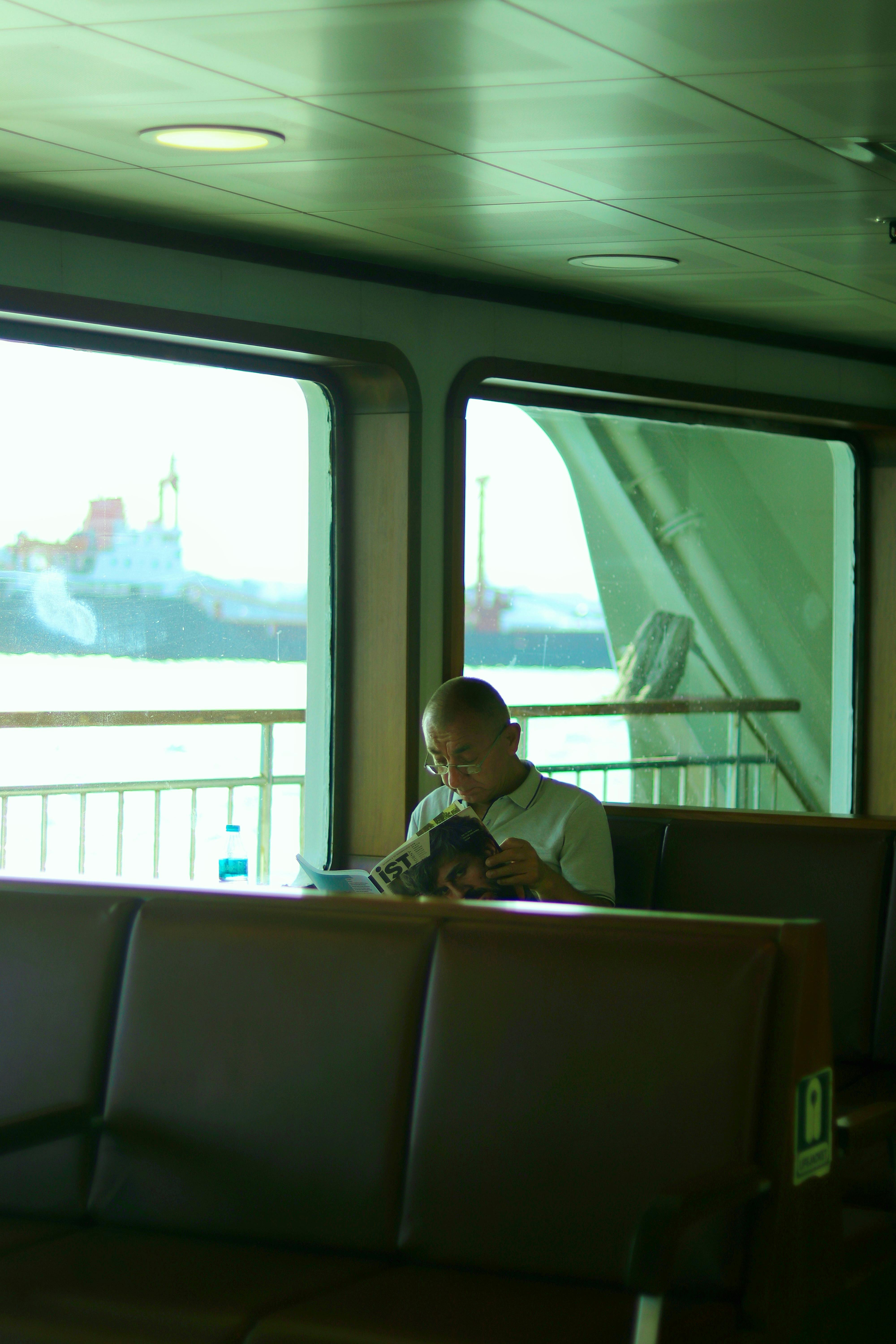 A man sits reading a magazine inside a passenger ferry on a sunny day.