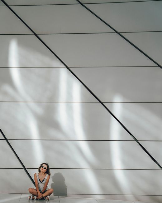 A woman with sunglasses sits against a sunlit, geometric modern wall casting artistic shadows.