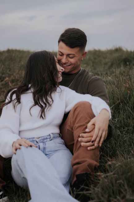 A loving couple sits closely on a meadow grass, sharing warm smiles and gentle moments.
