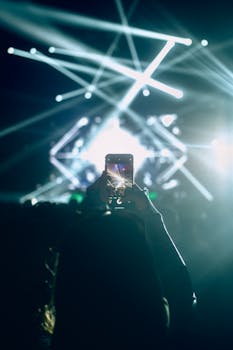 Vibrant concert scene with a crowd capturing moments on smartphones, illuminated by bright lights.