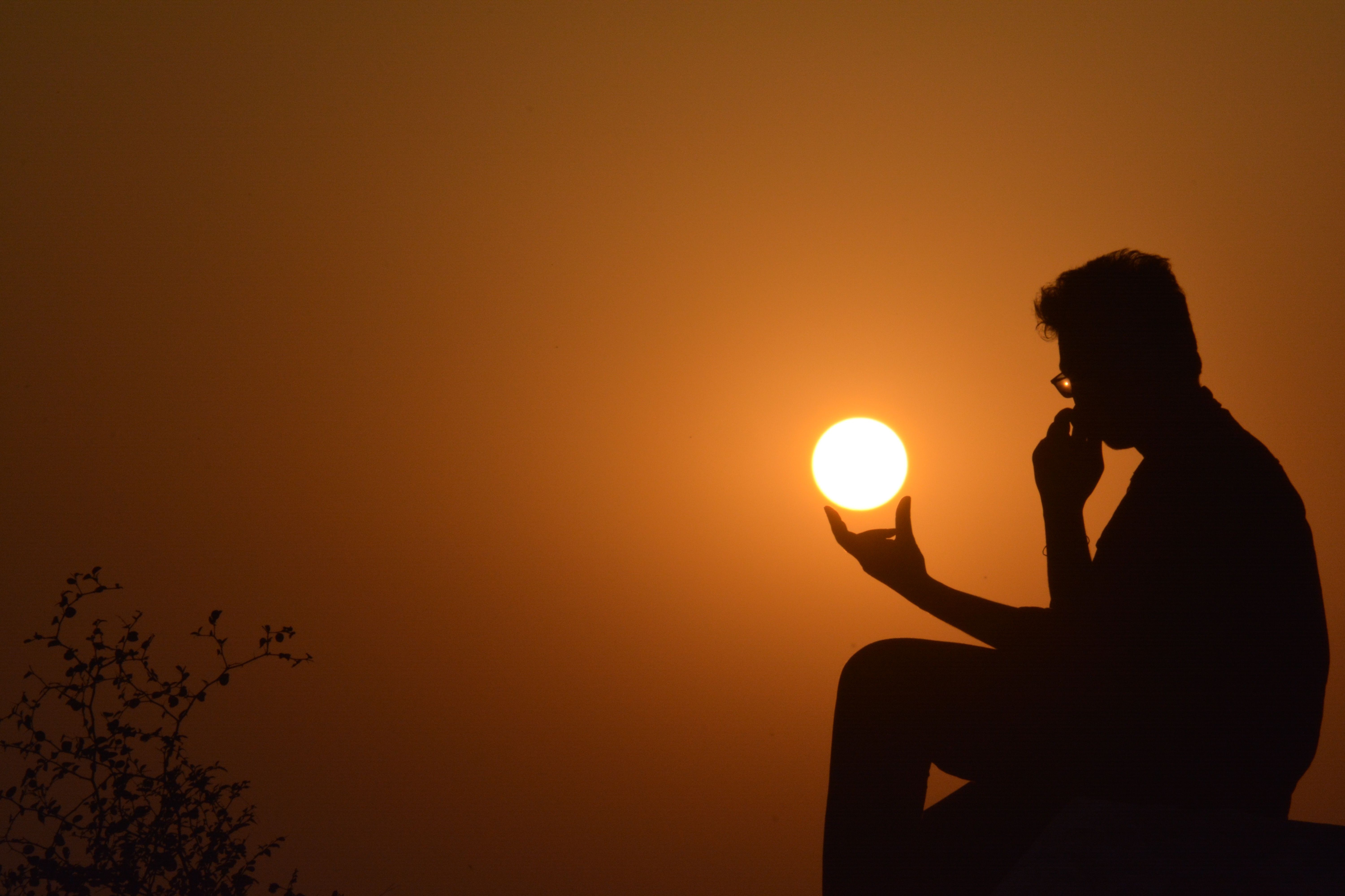 A silhouette of a man sitting at sunset, appearing to hold the sun with his hand.