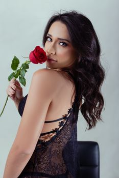 Young woman in lace lingerie holding a red rose, posing elegantly in a studio setting.
