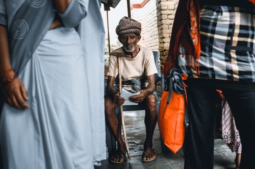 Free Senior man in traditional attire sitting with a walking stick in an urban street setting, surrounded by people. Stock Photo