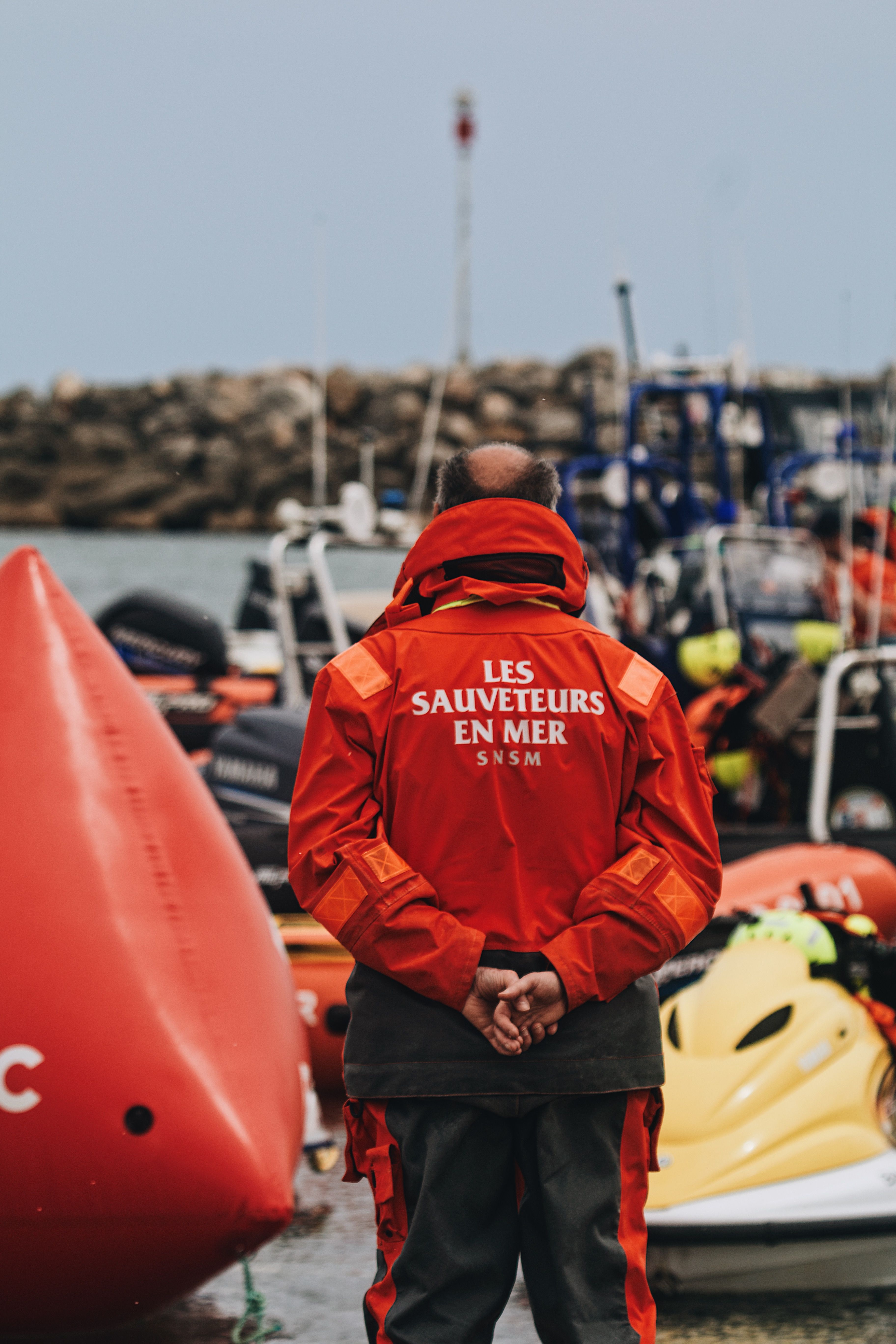 Lifeguard in red jacket standing among boats at a harbor, focused on safety and rescue.