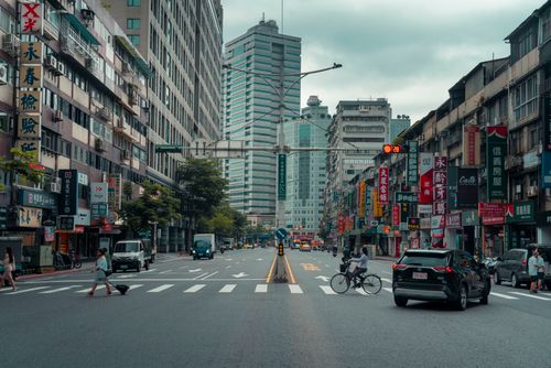 Free Bustling urban street scene capturing people, vehicles, and towering buildings under a cloudy sky. Stock Photo