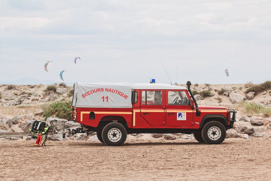 Red rescue truck parked on sandy beach with kite surfers in background, Secours Nautique visible.