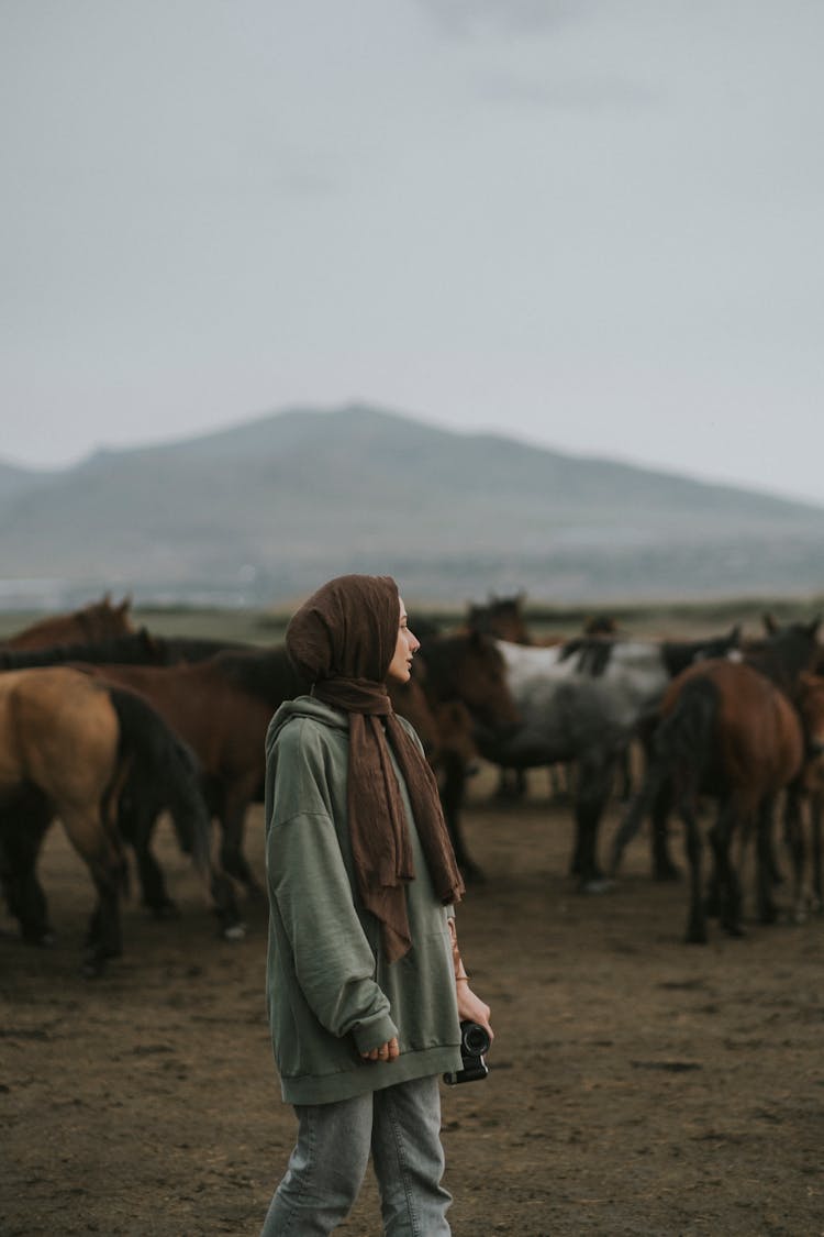 Young Woman With A Camera Standing On The Background Of An Herd Of Horses 