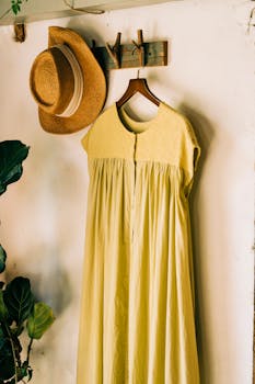 A light yellow dress and straw hat hanging on a rustic wall with green plants.