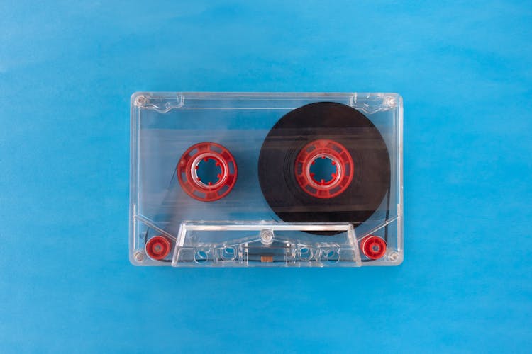 One Transparent Compact Audio Cassette With Visible Tape And Red Inner Reels Isolated On Blue Background. Top Down View Flat Lay With Empty Space For Text