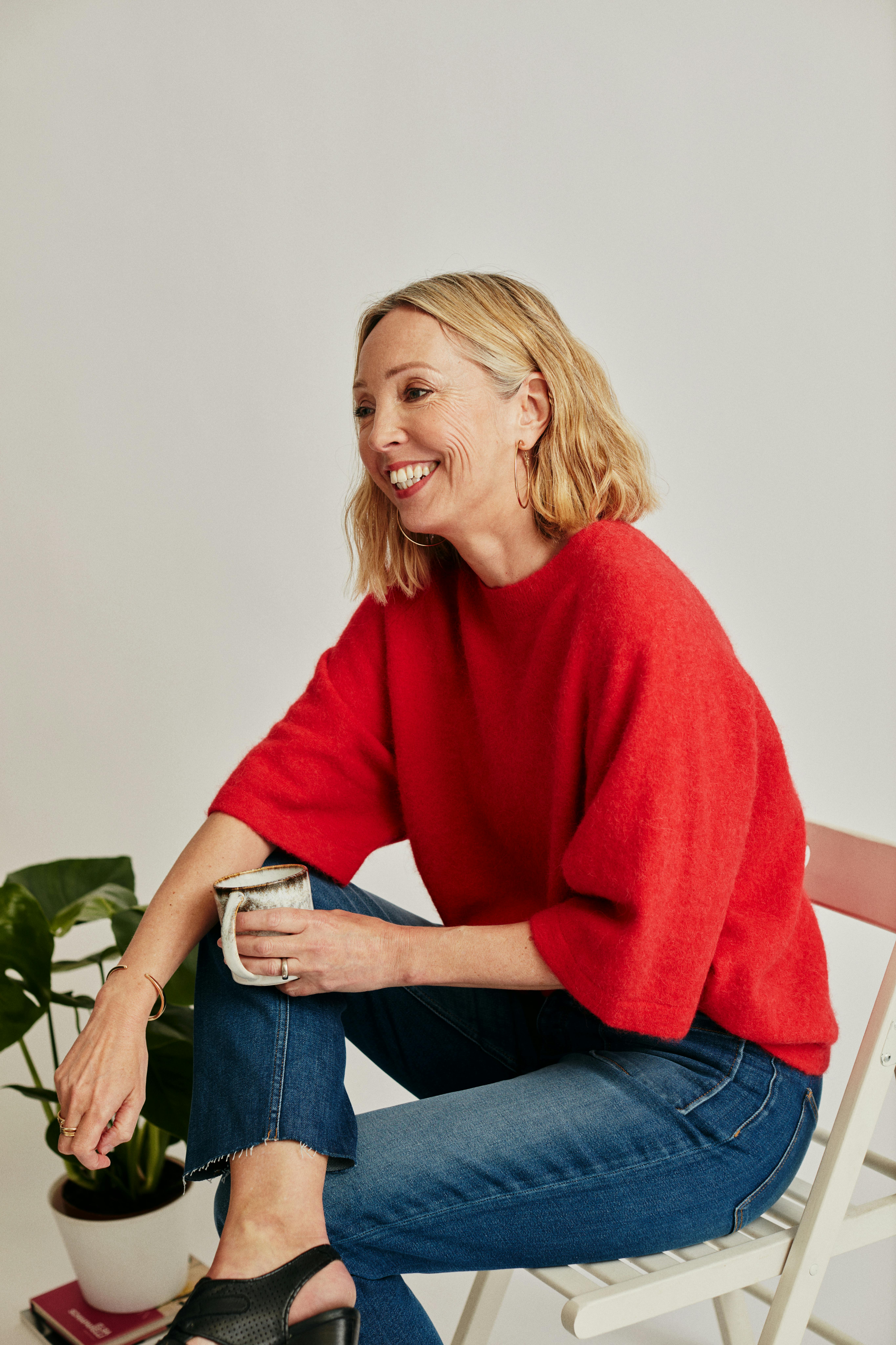 Stylish woman in red sweater sitting on chair, smiling in studio.