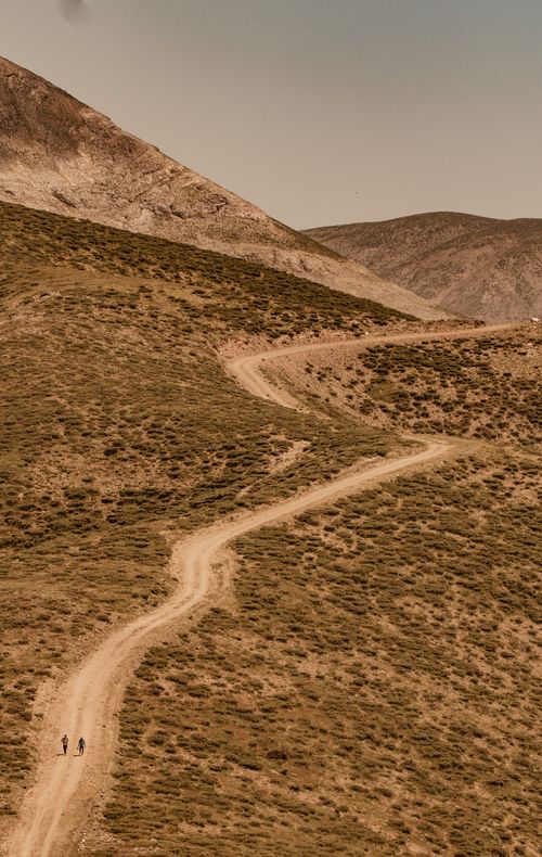 Free Drone view of a winding dirt road through dry, mountainous landscape with two people walking. Stock Photo
