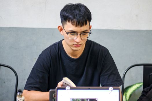 Teenager wearing eyeglasses reading a book at an indoor desk with a laptop.