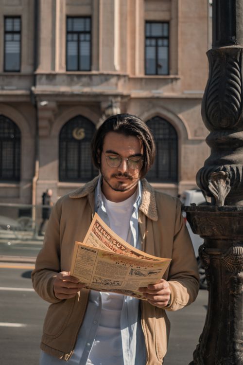 Free A man in fashionable attire reads a newspaper outdoors against an urban backdrop. Stock Photo