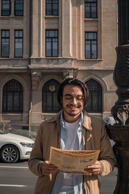 Free Young man in glasses reading a newspaper on a sunny day in the city. Casual fashion captured on an urban backdrop. Stock Photo