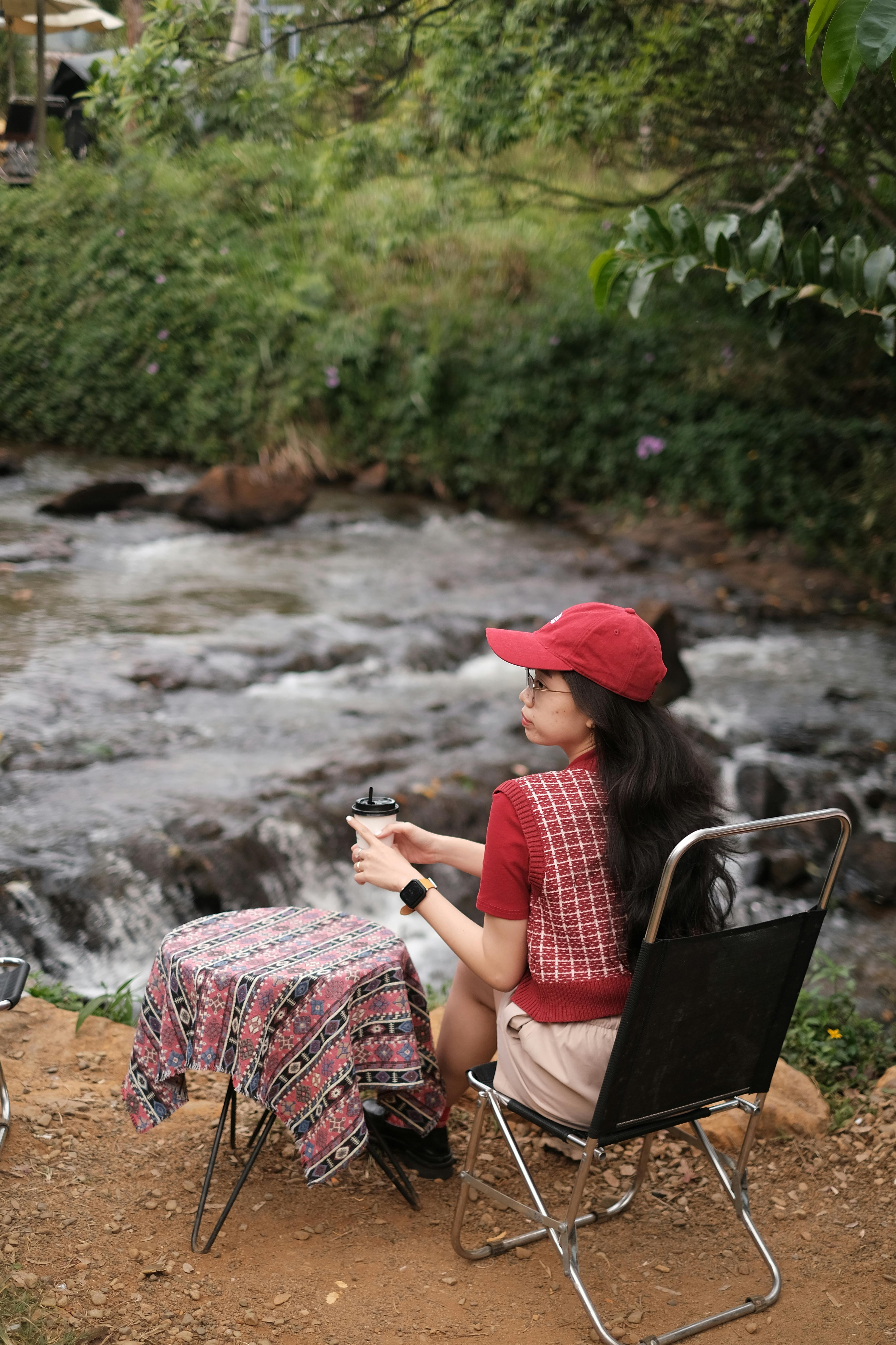 A woman relaxes by a riverside with a picnic setup, enjoying nature's tranquility.