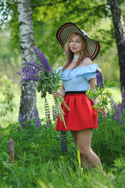Young Woman in a Summer Outfit Standing Outside and Holding a Bunch of Purple Lupins