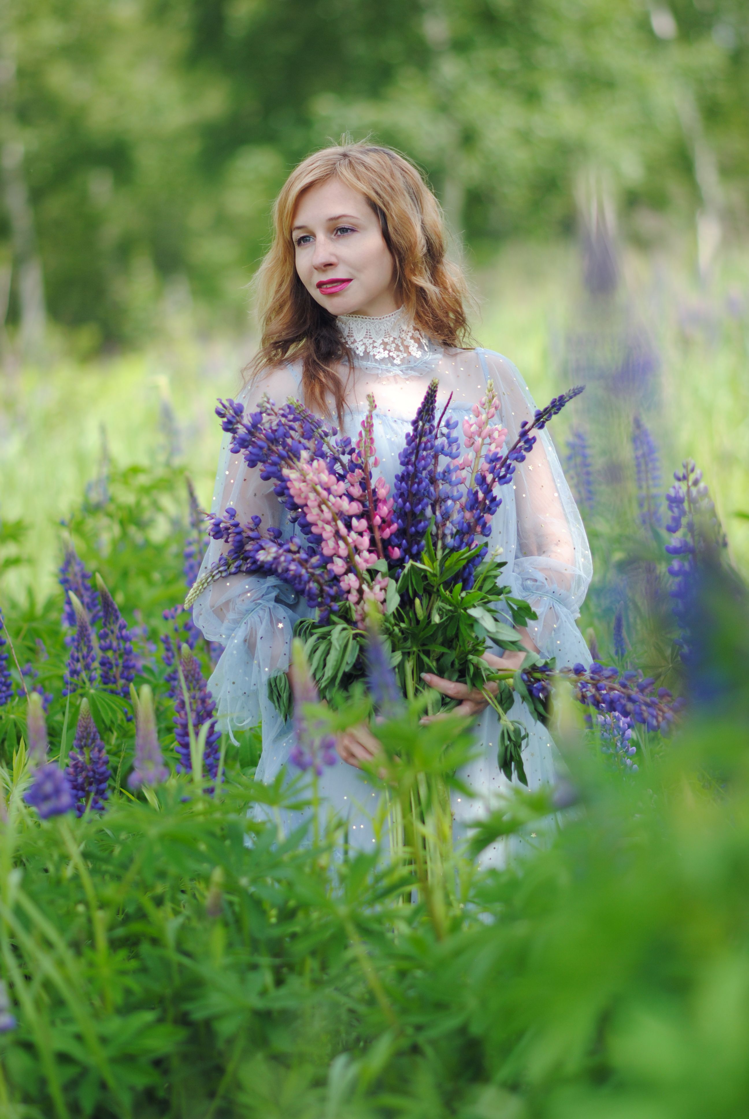 A young woman in a floral dress poses with a bouquet of lupins in a lush green field.