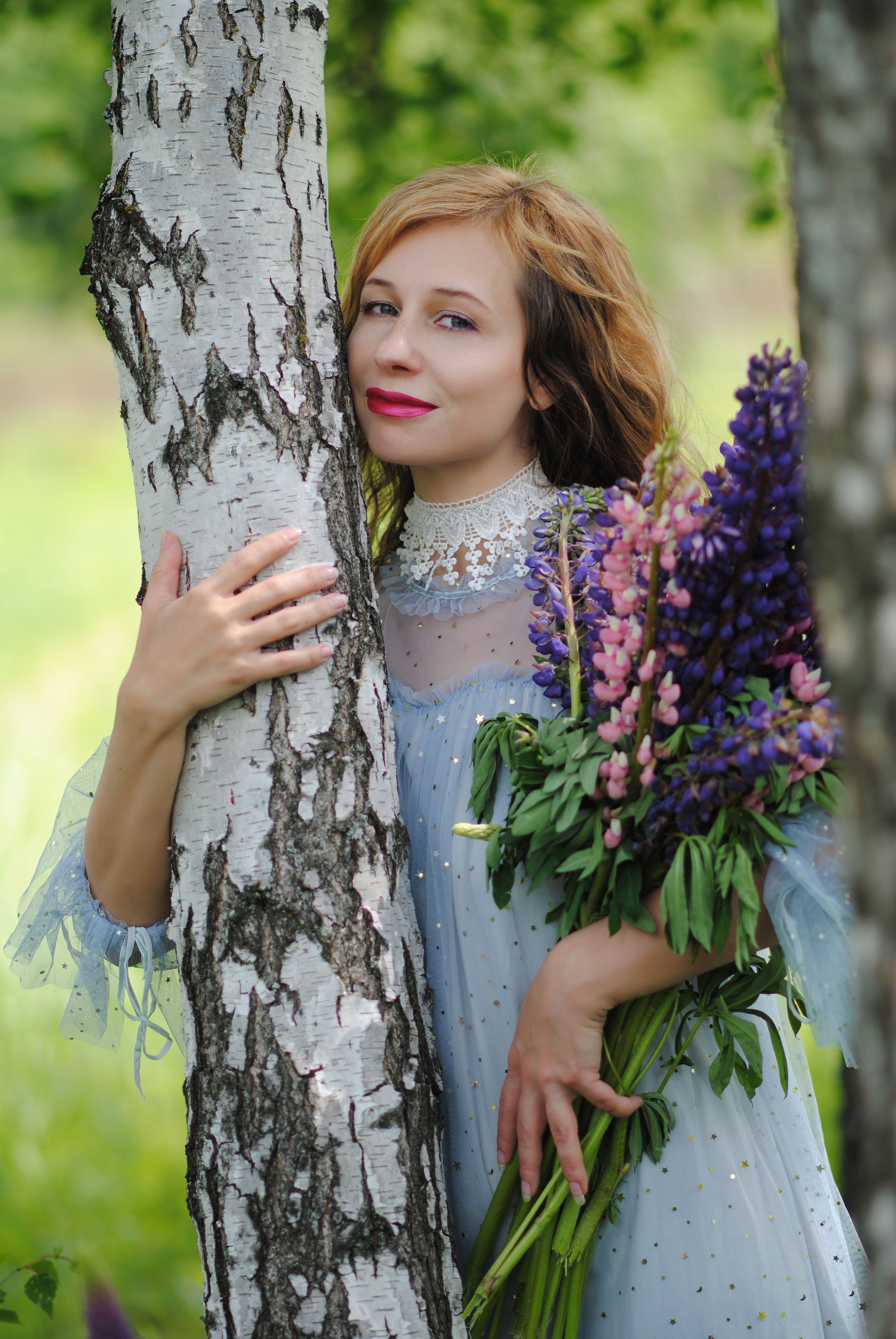A woman in a lace dress hugs a birch tree holding a beautiful lupine bouquet outdoors.