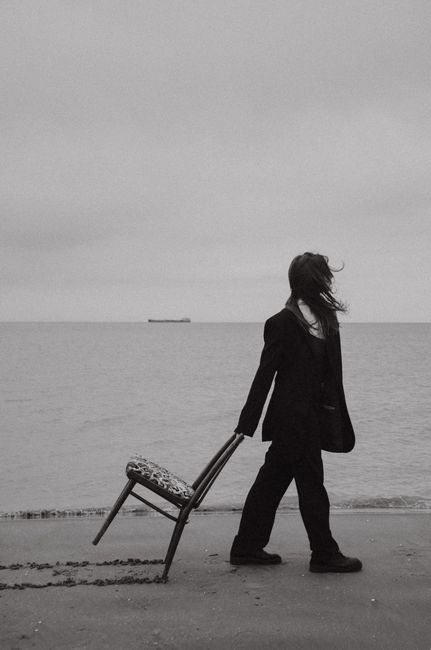 Artistic black and white photo of a woman dragging a chair on a tranquil beach.