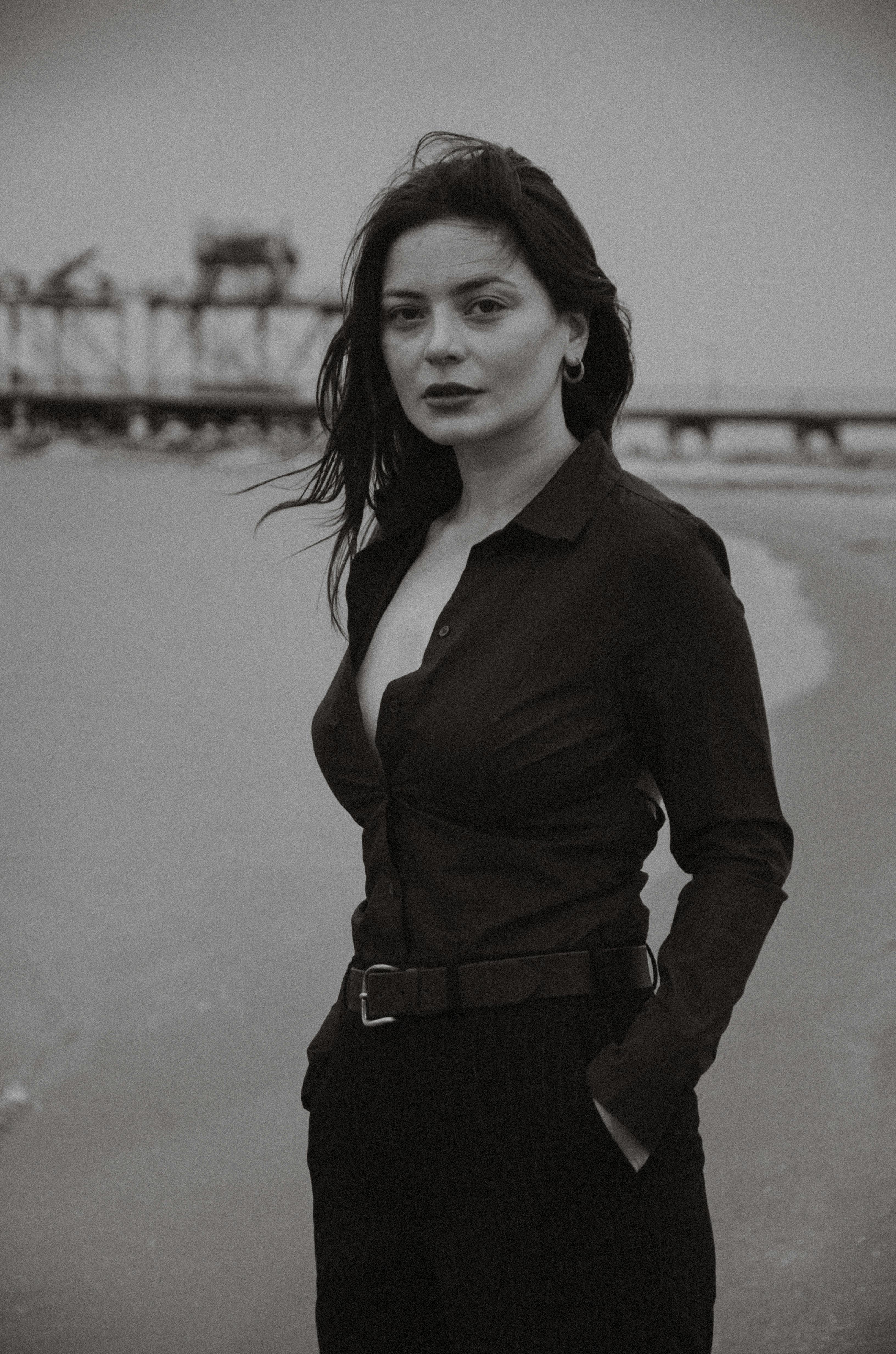 Black and white portrait of a stylish woman posing by the sea with a pier in the background.