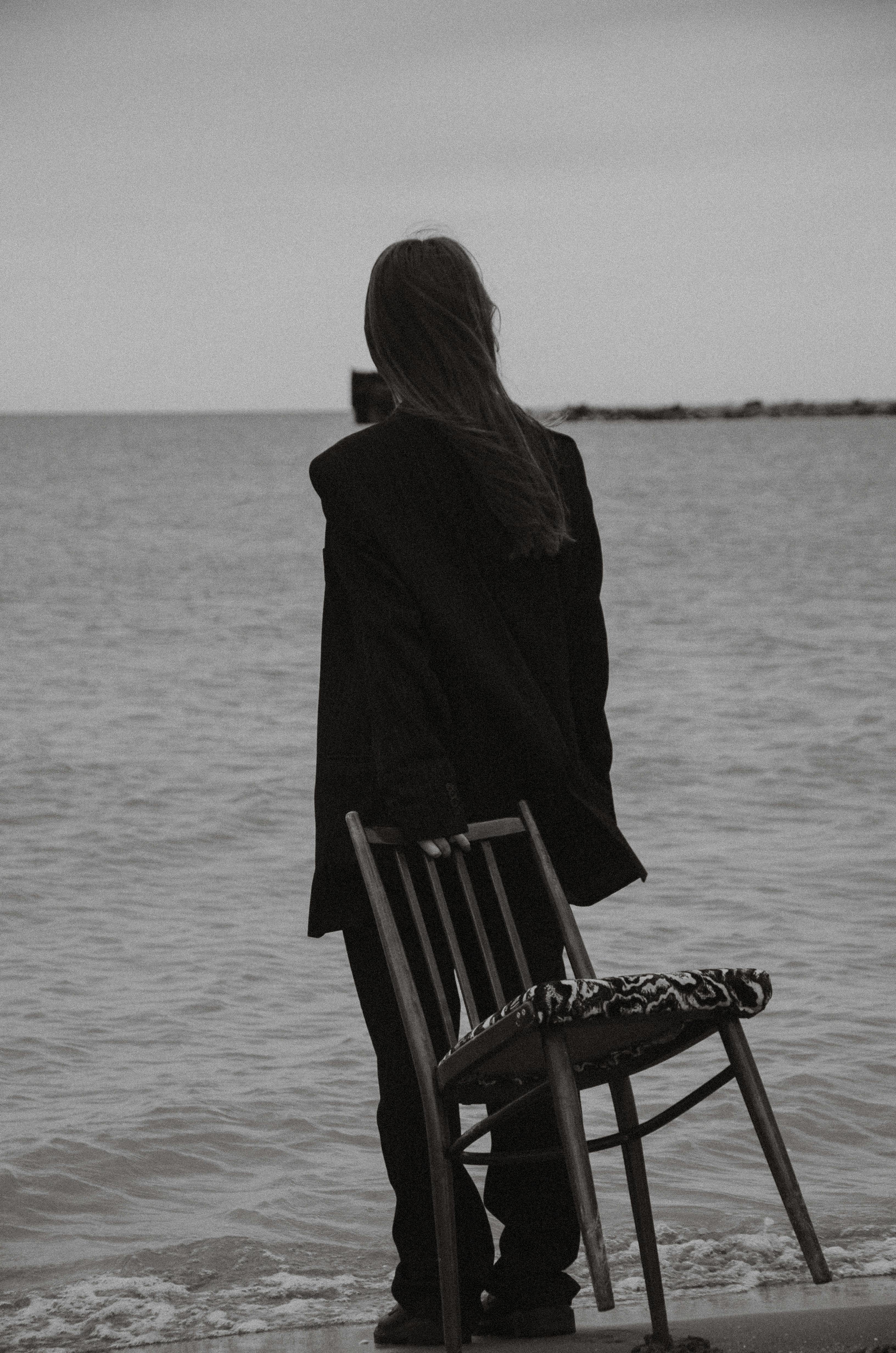 Black and white photo of a woman in contemplation by the ocean, holding a chair.