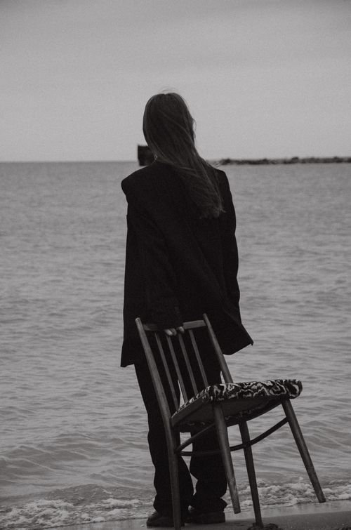 Free Black and white photo of a woman in contemplation by the ocean, holding a chair. Stock Photo