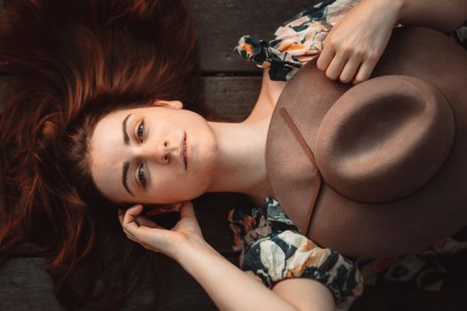 Artistic portrait of a woman lying on wooden floor holding a fedora hat, wearing a floral dress.