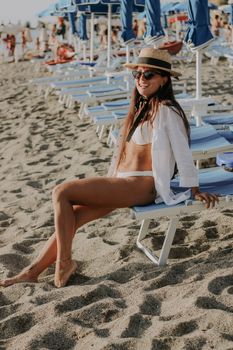 Woman in bikini and straw hat enjoying a sunny day at the beach, sitting on a sunbed.