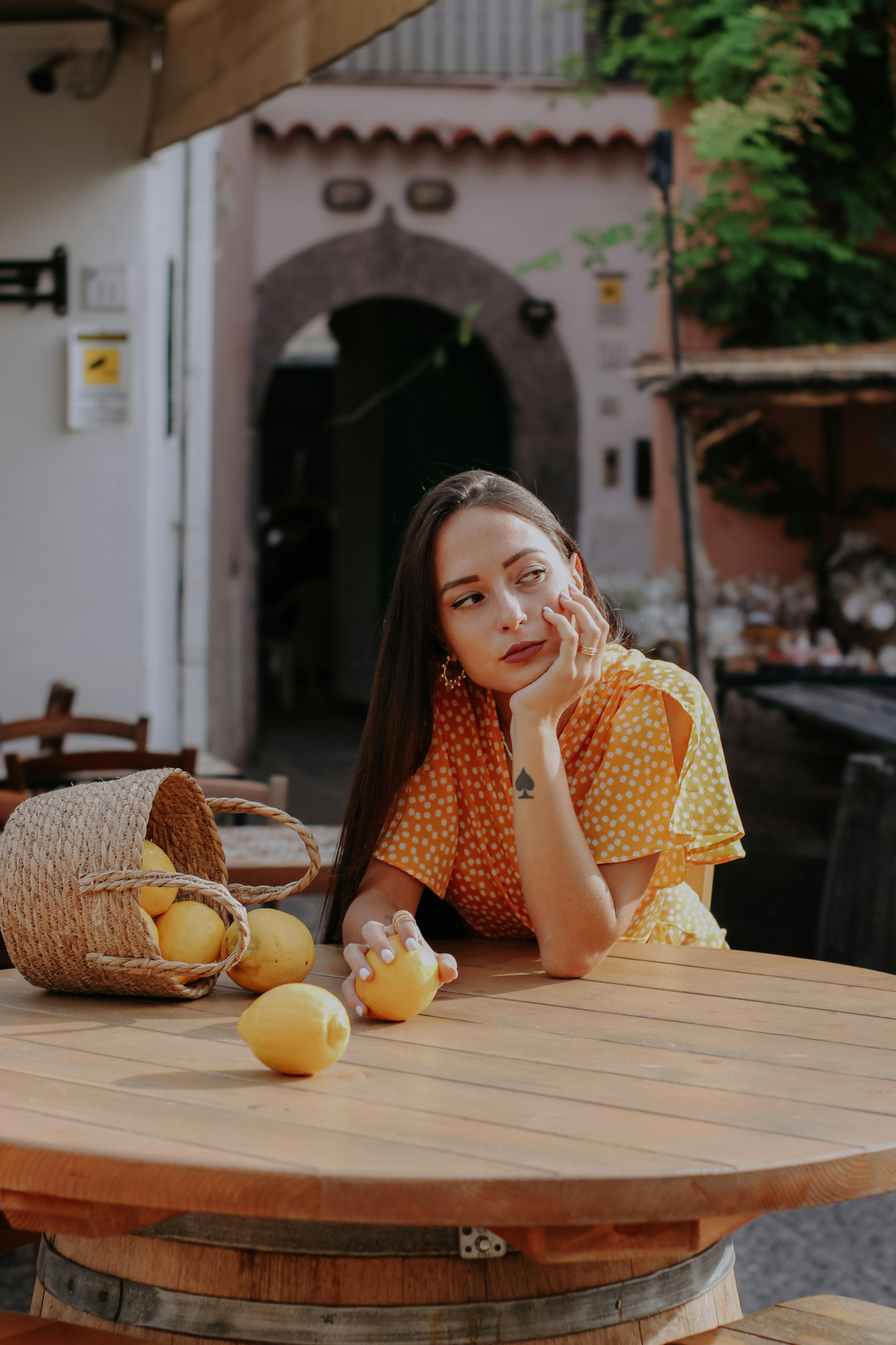 Woman in a yellow polka-dot dress sits thoughtfully at a table with lemons.