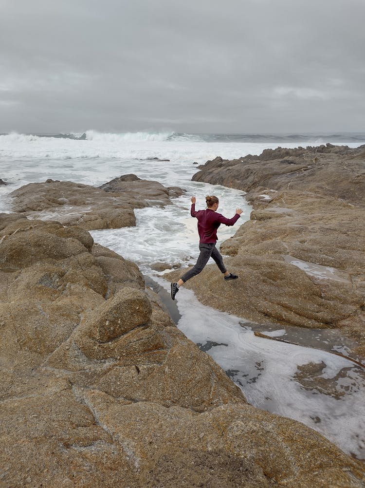 Woman Jumping Over Stream On Sea Shore Under Rain Clouds