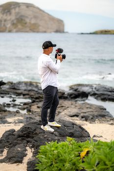 Male photographer standing on rocky beach, capturing ocean scenery with camera.