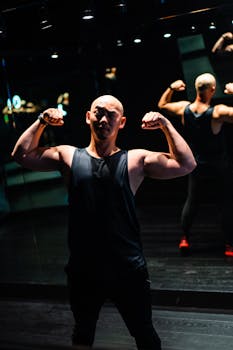 Bald man in a sleeveless top flexes muscles in a dimly lit gym with mirrors.