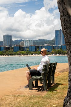 Man sitting on a bench by the sea with a laptop, urban skyline backdrop