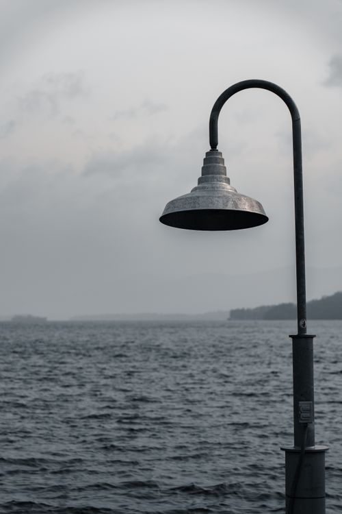 Free A lone metal street lamp stands against a monochrome seascape and cloudy sky, creating a serene yet dramatic scene. Stock Photo
