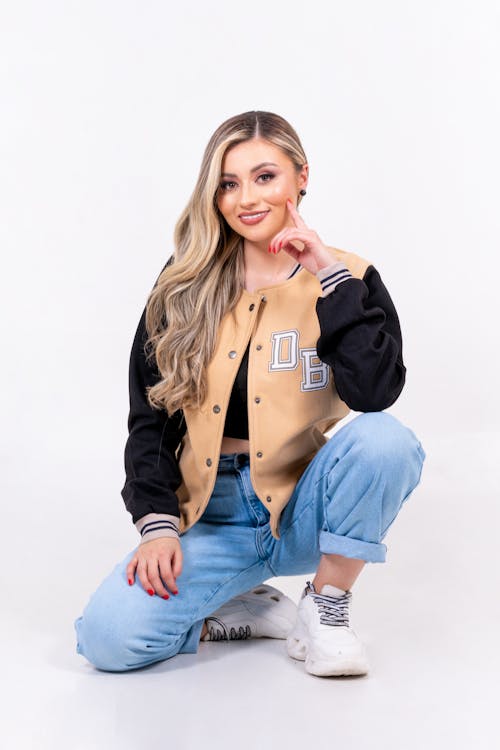Blonde Woman in Baseball Jacket and Jeans Crouching in Studio