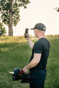 A man in a black t-shirt filming outdoors using a smartphone and professional camera.