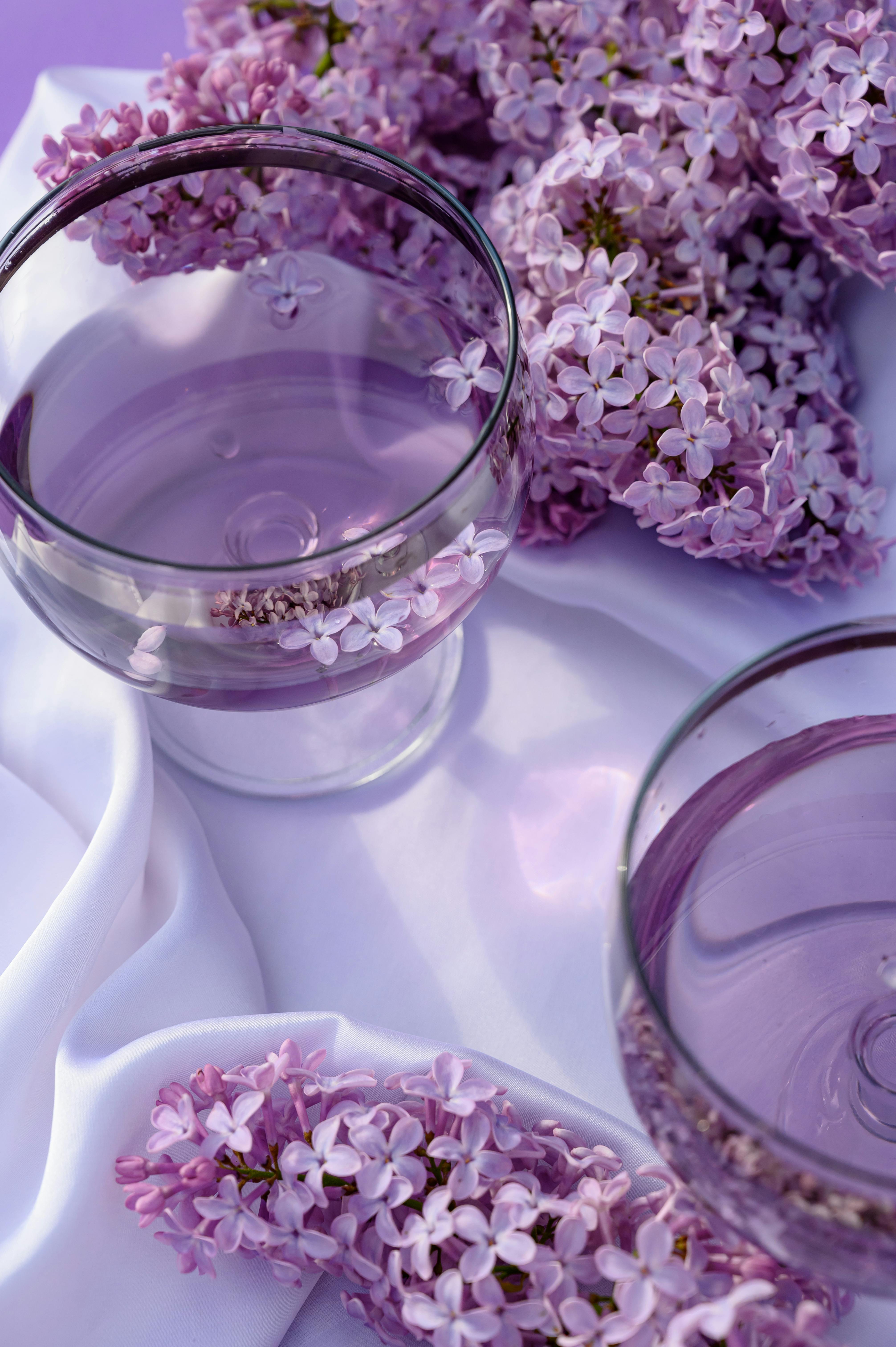 Lavender water in glass bowls surrounded by lilac flowers on a white cloth, evoking calmness.
