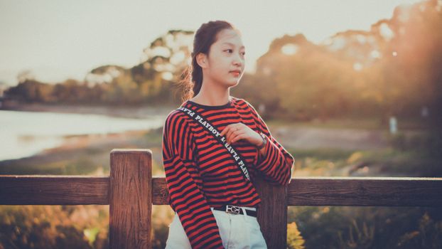 日落时分，一位身着条纹衬衫的年轻女子站在湖边栅栏旁，享受着温暖的阳光