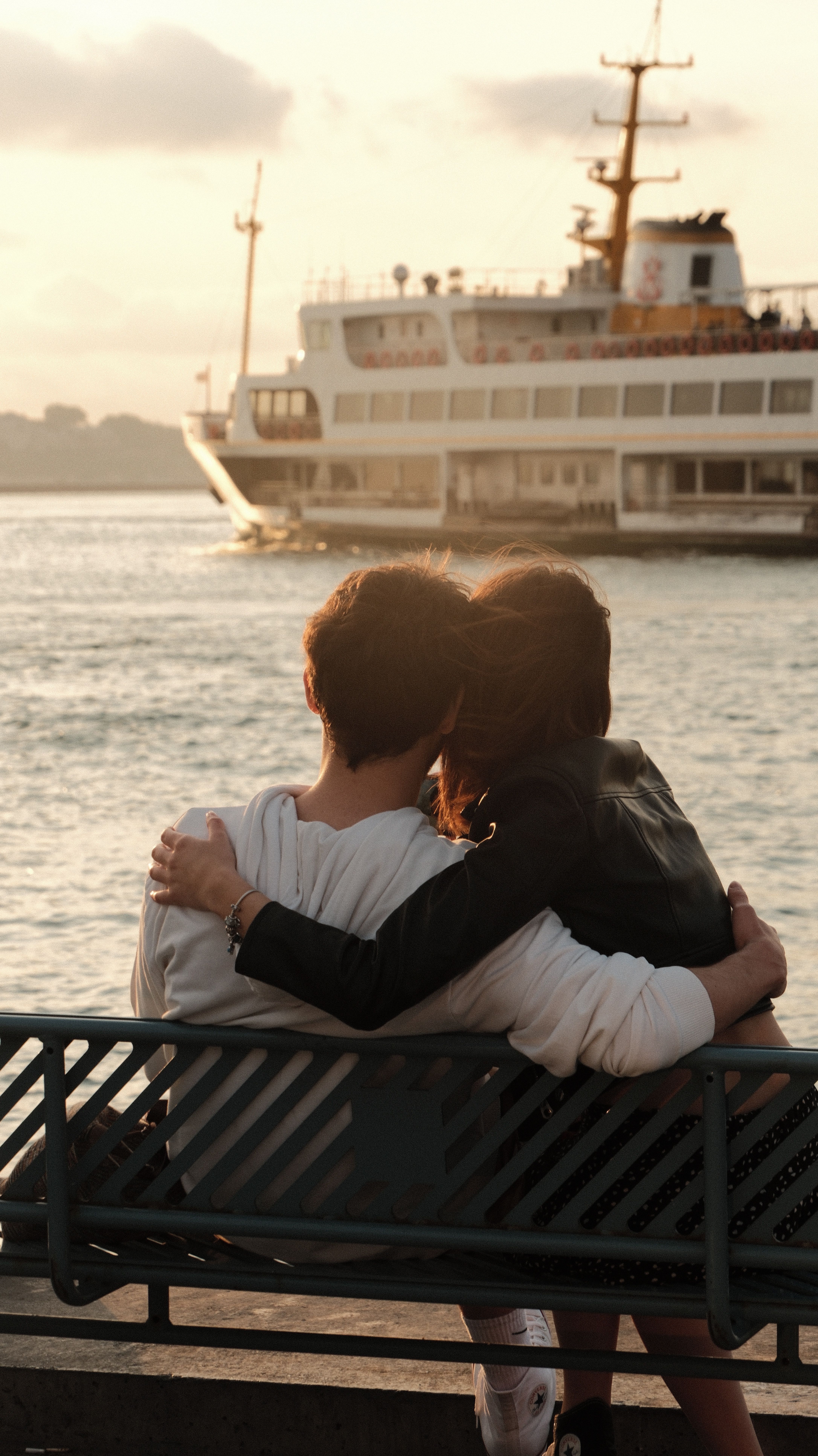 Teenage couple embraced on a bench by the sea, with a ferry passing in the background during sunset.