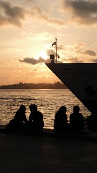 Silhouetted people sitting by a ship at sunset, overlooking Istanbul's skyline.