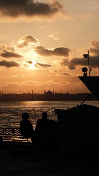 Silhouettes of people relaxing by the sea as the sun sets behind distant city skyline.