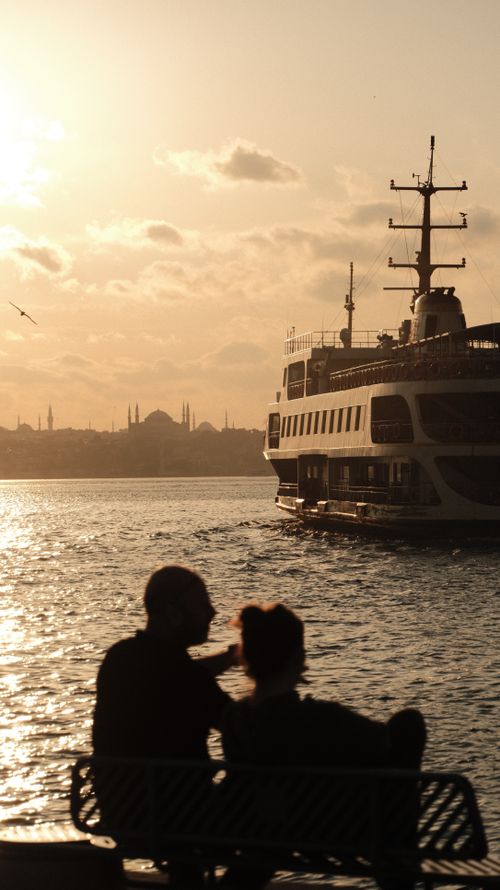 Free A tranquil evening view with a ship and people silhouetted on the Bosphorus, under a warm sunset. Stock Photo