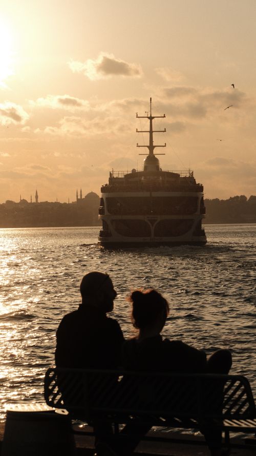 Free A couple watches a ferry silhouetted against the sunset on a serene evening by the sea. Stock Photo